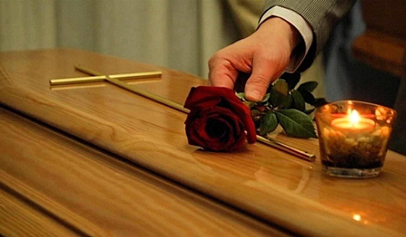 Una mano posa una rosa rossa sul feretro, accanto a una candela accesa